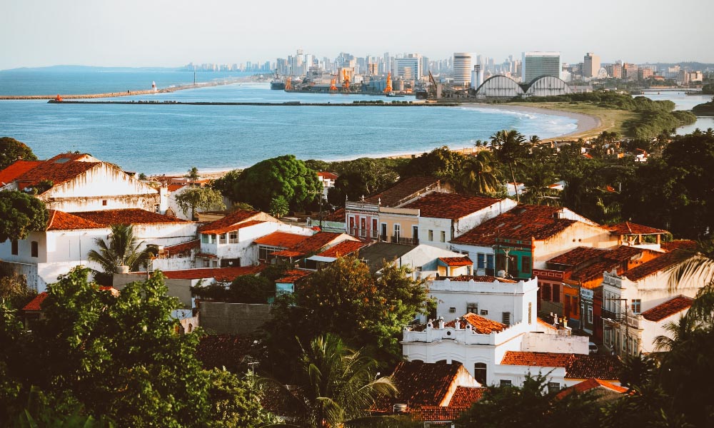 Vista de Olinda com casas coloridas, árvores e o mar ao fundo com prédios de Recife